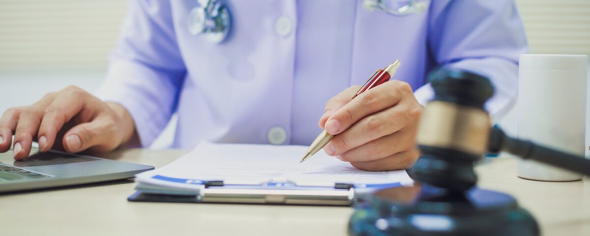 A close-up of the hands of a person wearing a white robe and a stethoscope behind a desk with a judge's gavel on it.