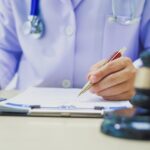 A close-up of the hands of a person wearing a white robe and a stethoscope behind a desk with a judge's gavel on it.