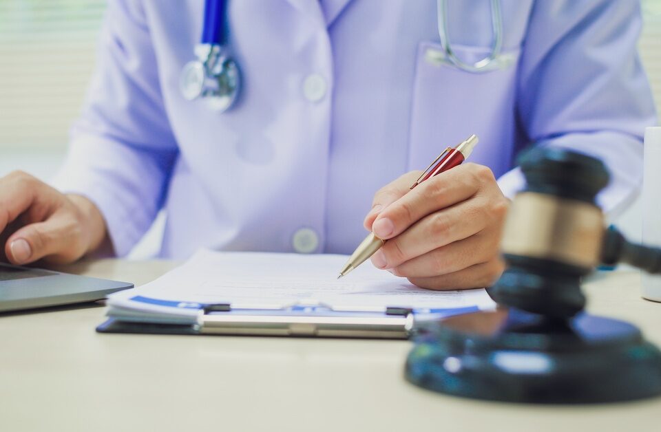 A close-up of the hands of a person wearing a white robe and a stethoscope behind a desk with a judge's gavel on it.