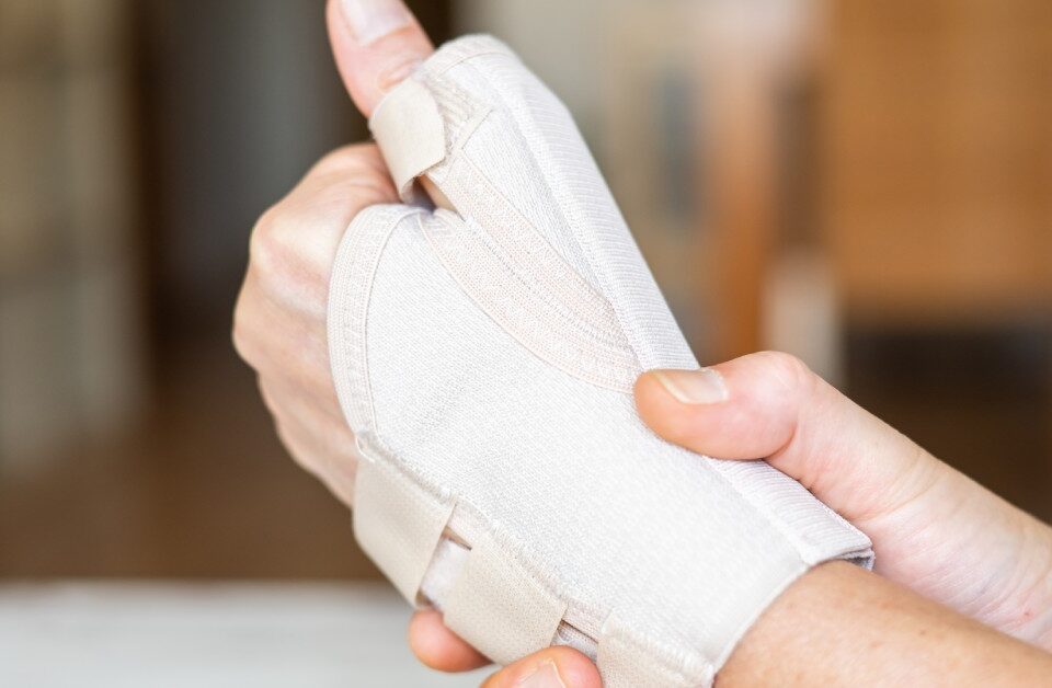A close-up of a hand wearing an orthopedic brace while holding the wrist with the other hand, with a blurry background.