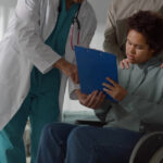 A young man in a wheelchair, holding and looking at a blue clipboard with a man in a white robe next to him.