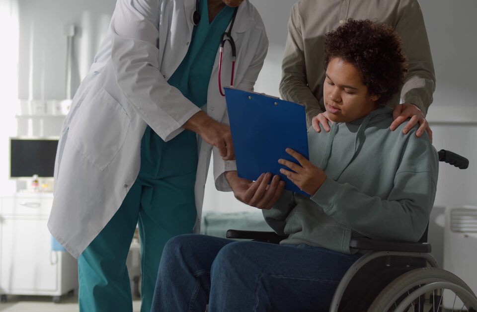 A young man in a wheelchair, holding and looking at a blue clipboard with a man in a white robe next to him.
