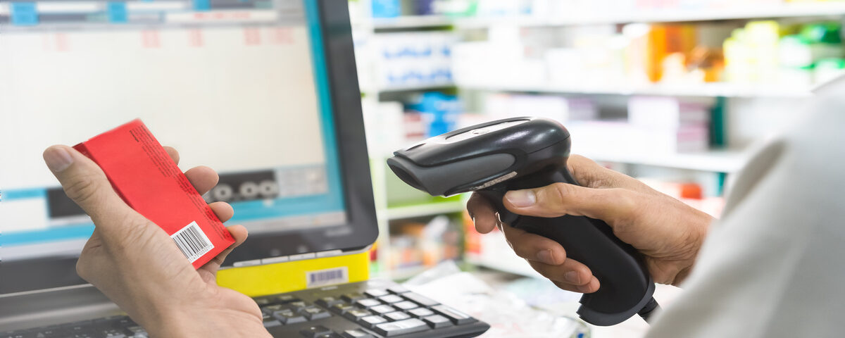 Two hands holding a small box and a scanner inside a pharmacy with multiple medicine boxes in the background.