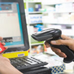 Two hands holding a small box and a scanner inside a pharmacy with multiple medicine boxes in the background.