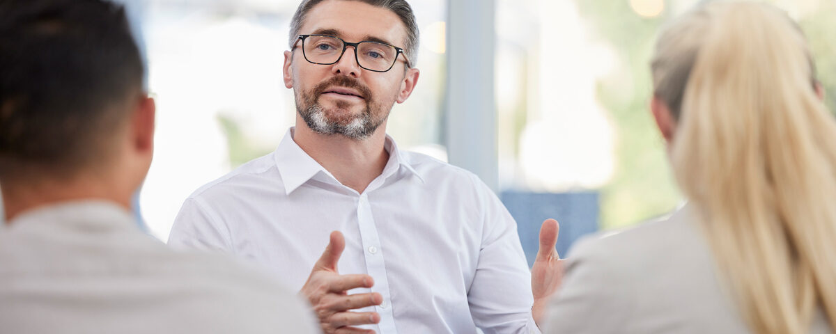 A man with a grey beard, wearing a white shirt and glasses, sitting across from a blurred man and a woman.