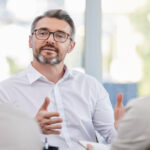 A man with a grey beard, wearing a white shirt and glasses, sitting across from a blurred man and a woman.