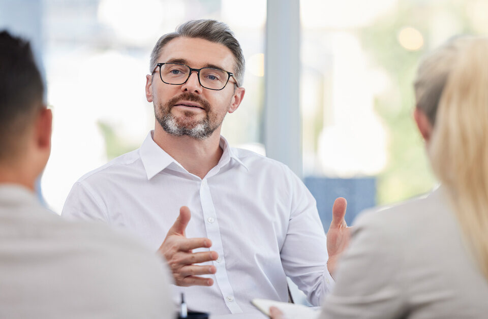 A man with a grey beard, wearing a white shirt and glasses, sitting across from a blurred man and a woman.